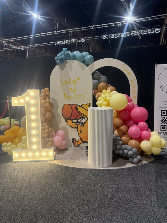 A custom 1st birthday party backdrop setup featuring a large illuminated marquee number 1, a white arched sign reading 'Sweet as honey' with Winnie the Pooh illustration, a white ribbed cylinder pillar, and balloon garlands in pink, yellow, brown, and gray.