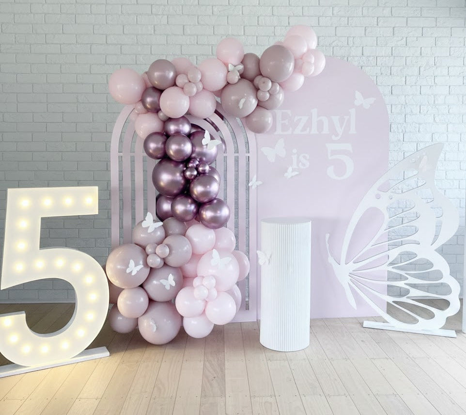 Balloon arch with pink and purple balloons against a white brick wall with decorative elements.