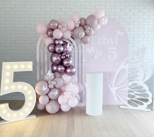 Balloon arch with pink and purple balloons against a white brick wall with decorative elements.